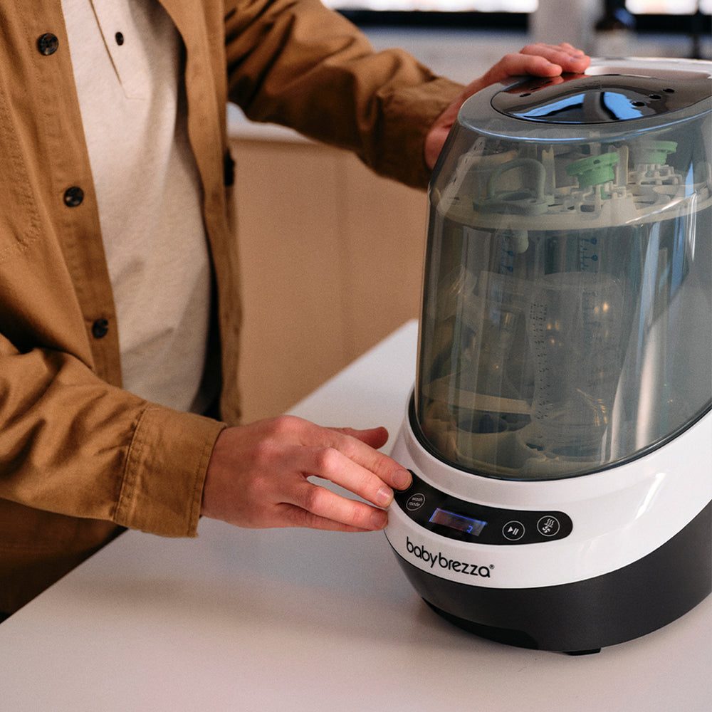 A person in a brown jacket is using a Baby Brezza bottle washer pro on a white countertop in a kitchen setting.
