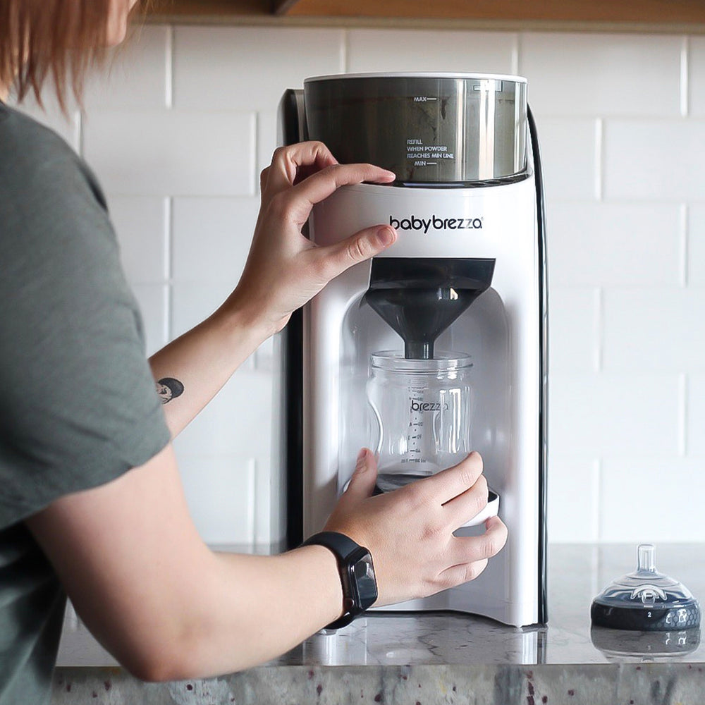 A person inserts a clear baby bottle into a silver and black Baby Brezza formula pro advanced machine on a kitchen counter.