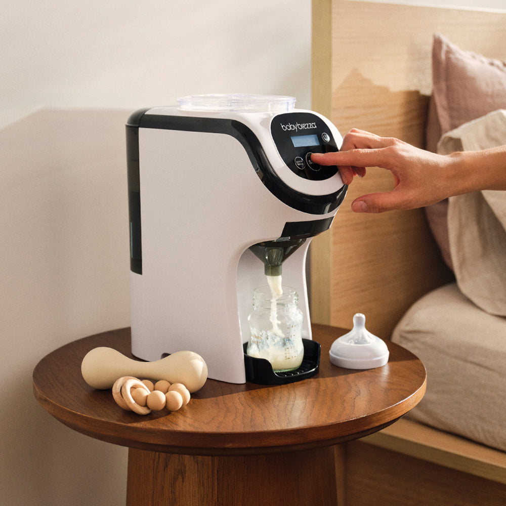 A Baby Brezza formula pro mini machine on a wooden side table, with a hand pressing the control panel. A baby bottle and bottle cap sit nearby, with a wooden teething toy in the foreground, set in a bedroom environment.