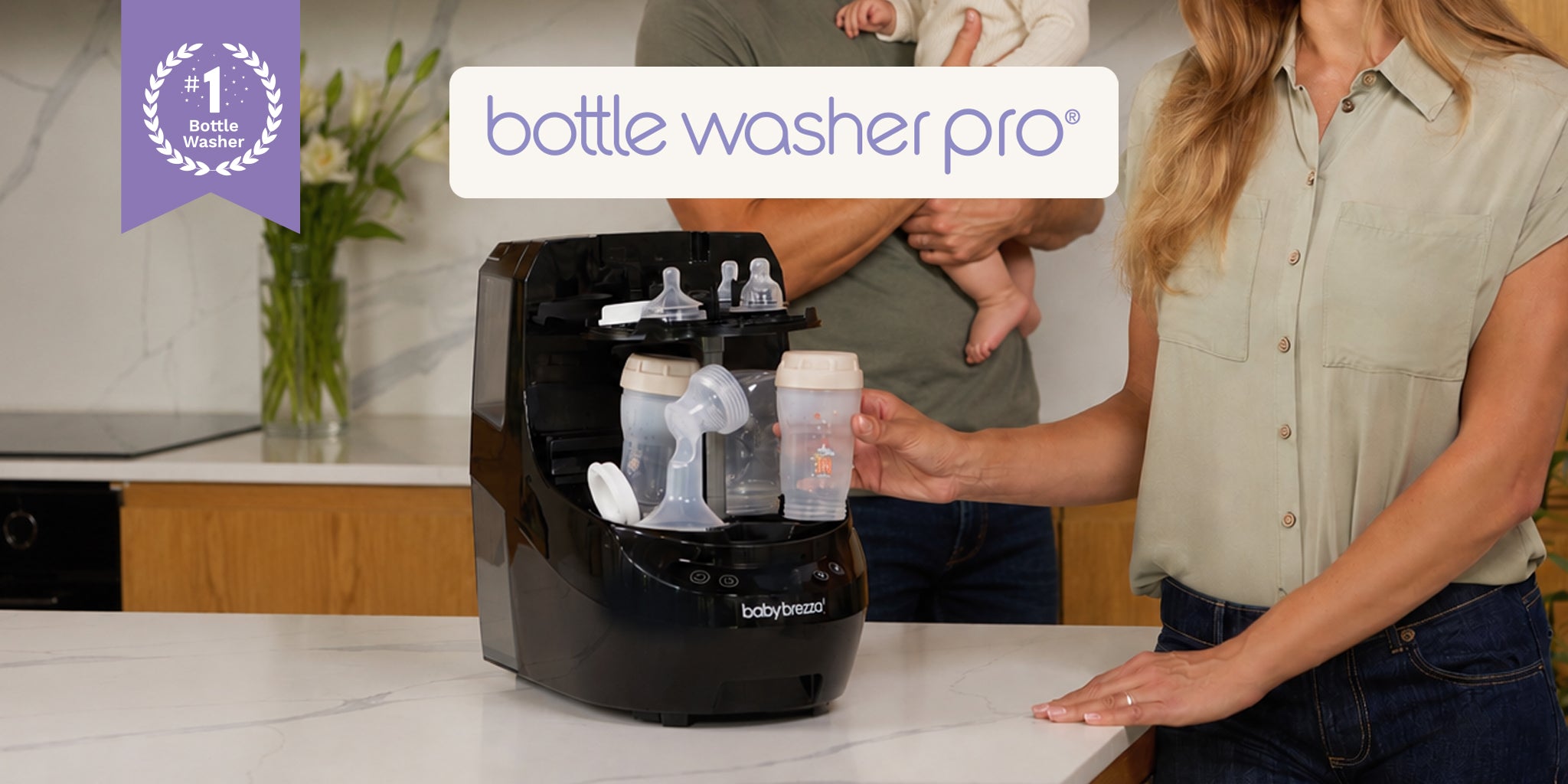 A Baby Brezza bottle washer pro on a kitchen counter with a woman standing beside it and a baby being held in the background. A purple badge in the top left says 'number 1 bottle washer' and a white banner at the top reads 'bottle washer pro'.