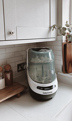 A Baby Brezza bottle washer pro, placed on a white kitchen countertop against a white subway tile backsplash.