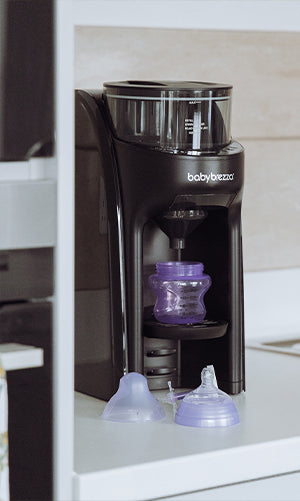 A Baby Brezza Formula Pro Advanced machine in black, with a purple baby bottle beneath the dispenser. Two purple bottle caps are placed on the white countertop in front of the machine.