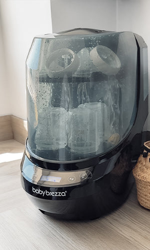 A Baby Brezza bottle washer pro, shown with baby bottles inside on a light wood floor.