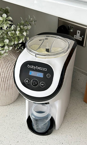 A Baby Brezza formula pro machine in white and black, shown on a speckled kitchen countertop with a baby bottle placed underneath.