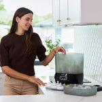 A woman smiles in a modern kitchen with white cabinets, using the Baby Brezza UK Steriliser Dryer Advanced with HEPA filter on the counter. Clean bottle parts are nearby, and a tiled backsplash adds to the bright space. - product thumbnail
