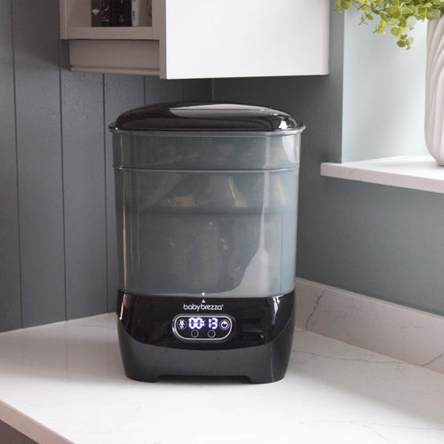 A black and clear Baby Brezza UK Steriliser Dryer Advanced with a HEPA filter sits on a white countertop beneath a window, its display showing 13 minutes. Plants and kitchen cabinets are visible in the background. - product thumbnail