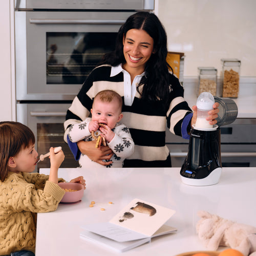 A smiling woman holds a baby and prepares a bottle with the Baby Brezza UK Bottle & Breastmilk Warmer in a modern kitchen. A child eats cereal nearby, with an open book and plush toy on the white countertop. - product thumbnail