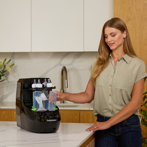 A woman in a kitchen places a baby bottle into the black Baby Brezza UK Bottle Washer Pro—an all-in-one washer, steriliser, and dryer—on the countertop. - product thumbnail