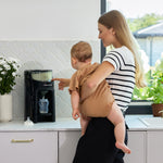 A woman holding a baby uses her free hand to operate the Baby Brezza BUNDLE Formula Pro Advanced And SuperQuick Steriliser (All Black) by Baby Brezza UK on the kitchen counter, with a baby bottle below, flowers and a window in the background. - product thumbnail