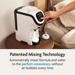 A user presses a button on the Baby Brezza UK BUNDLE Formula Pro Mini White preparing formula on a wooden table. Text below highlights its patented mixing tech for smooth, air bubble-free formula and features the SuperQuick Steriliser Black. - product thumbnail