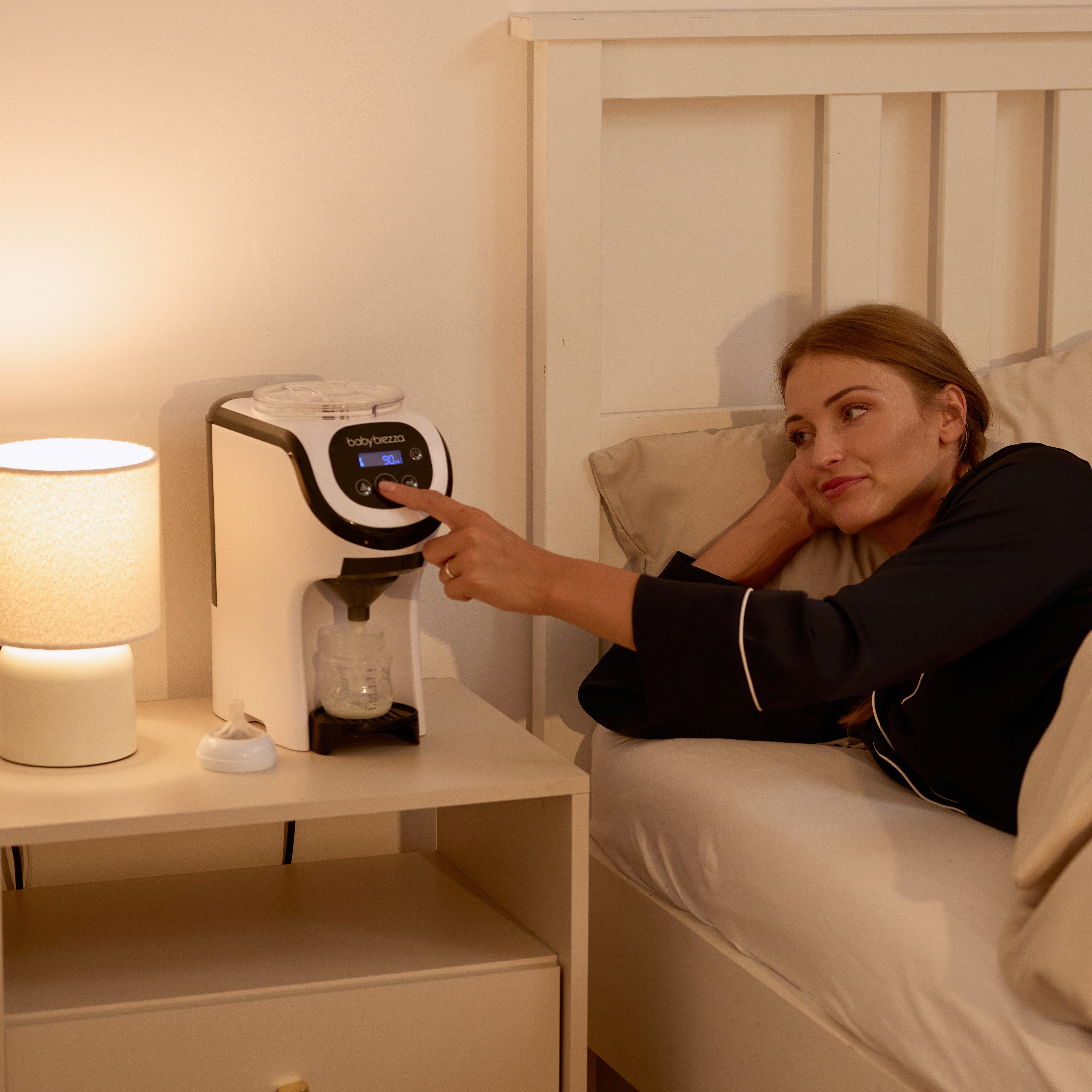A woman smiles at a young girl sitting on a bed near a crib in a cozy bedroom with large windows. A Baby Brezza UK Formula Pro Mini automatic formula maker is on a small table by the bed. Snow-covered trees are visible outside. - product thumbnail