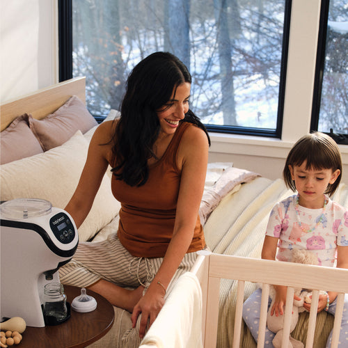A woman smiles at a young girl sitting on a bed near a crib in a cozy bedroom with large windows. A Baby Brezza UK Formula Pro Mini automatic formula maker is on a small table by the bed. Snow-covered trees are visible outside. - product thumbnail