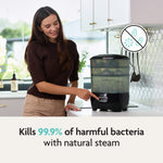 In a modern kitchen, a woman touches the Baby Brezza UK BUNDLE Steriliser Dryer Advanced Black and Bottle & Breast Milk Warmer. Text reads, “Kills 99.9% of harmful bacteria with natural steam” with a crossed-out bacteria graphic. - product thumbnail