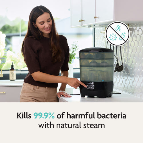 In a modern kitchen, a woman touches the Baby Brezza UK BUNDLE Steriliser Dryer Advanced Black and Bottle & Breast Milk Warmer. Text reads, “Kills 99.9% of harmful bacteria with natural steam” with a crossed-out bacteria graphic. - product thumbnail
