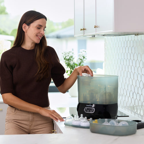 A woman stands in a kitchen placing baby bottles into a Baby Brezza BUNDLE Steriliser Dryer Advanced Black and Bottle & Breast Milk Warmer by Baby Brezza UK, with clean bottle parts arranged on the counter beside her. - product thumbnail