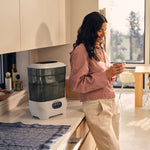 A woman with long hair stands smiling in a sunlit kitchen, holding a cup. Beside her on the counter is the Baby Brezza UK Steriliser Dryer Advanced with HEPA filter and a towel. Large windows reveal a blurred view outside. - product thumbnail