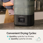 A person stands behind the Baby Brezza UK BUNDLE SuperQuick Steriliser Dryer & SuperFast Portable Bottle Warmer on a kitchen counter. Text highlights QuickDry (5 or 10 min) and QuietDry (25 min) cycles with a clock icon for fast drying. - product thumbnail