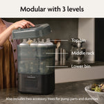 A person lifts the lid of the Baby Brezza UK BUNDLE Formula Pro Advanced And SuperQuick Steriliser - All Black on a kitchen counter beside an automatic formula maker. Labels show top bin, middle rack, lower bin, and accessory trees for pump parts and dummies. - product thumbnail