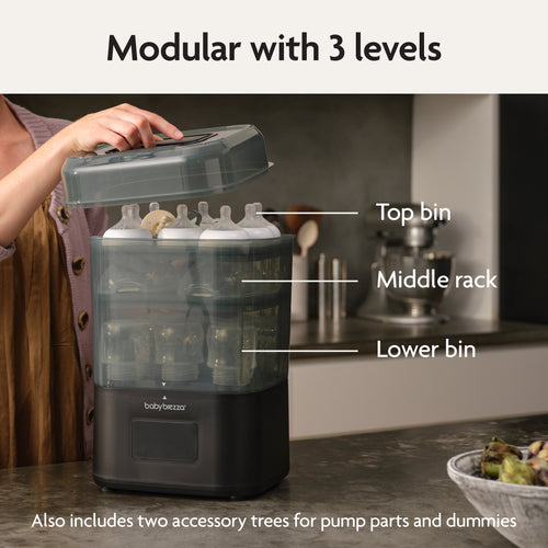 A person lifts the lid of the Baby Brezza UK BUNDLE Formula Pro Advanced And SuperQuick Steriliser - All Black on a kitchen counter beside an automatic formula maker. Labels show top bin, middle rack, lower bin, and accessory trees for pump parts and dummies. - product thumbnail