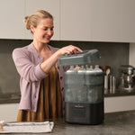 A woman in a kitchen, smiling in a light purple cardigan and brown dress, lifts the lid of the Baby Brezza UK BUNDLE Formula Pro Mini White and SuperQuick Steriliser Black filled with bottles; towels and utensils are on the counter nearby. - product thumbnail