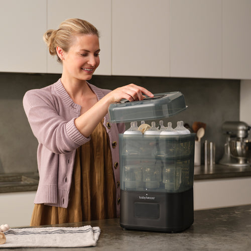 A woman smiles as she places a lid on the Baby Brezza BUNDLE SuperQuick Steriliser Dryer and SuperFast Portable Bottle Warmer by Baby Brezza UK, filled with baby bottles, in a modern kitchen. - product thumbnail