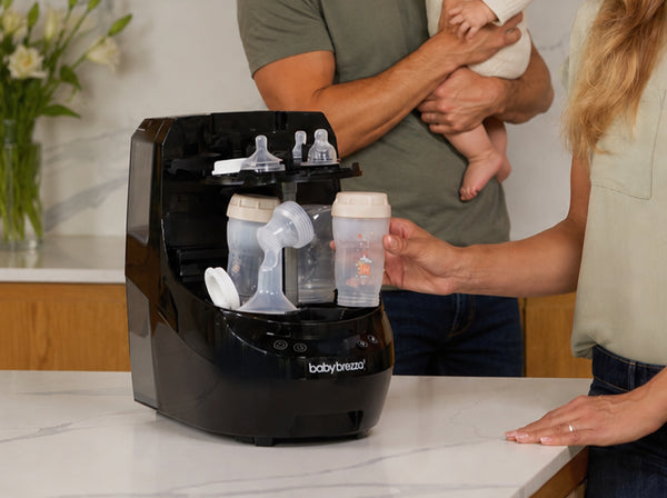 A black Baby Brezza Bottle Washer pro sits on a white marble countertop in a kitchen setting, with parents and a baby in the background.