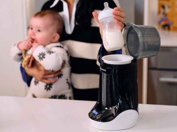 A Baby Brezza bottle + breastmilk warmer in black and white sits on a white countertop. A person withdraws a bottle of milk from the open container. A baby in a white patterned outfit is held in the background by the adult.