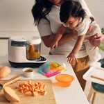A woman holds a baby in a kitchen with chopped sweet potatoes, the Baby Brezza UK One Step Food Maker Deluxe + FREE Pouches, pureed food, a colorful pouch, small bowl on the counter, and a high chair—ideal for homemade baby food. - product thumbnail