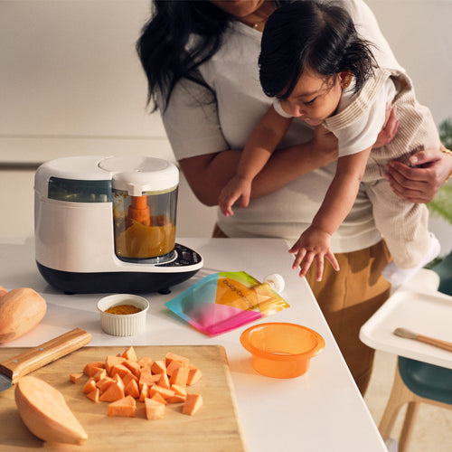 A woman holds a baby in a kitchen with chopped sweet potatoes, the Baby Brezza UK One Step Food Maker Deluxe + FREE Pouches, pureed food, a colorful pouch, small bowl on the counter, and a high chair—ideal for homemade baby food. - product thumbnail