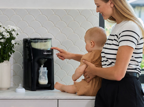 A mother in a black and white top holds a baby while using a Baby Brezza Formula Pro Advanced