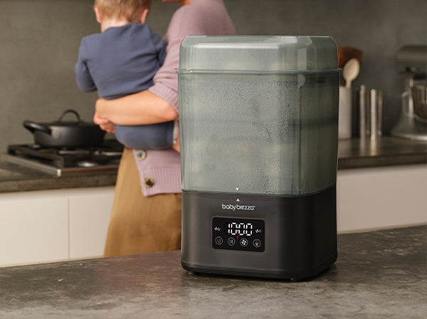 A Baby Brezza superquick 10-minute steriliser dryer sits on a kitchen countertop, with a blurred mother holding her child in the background.